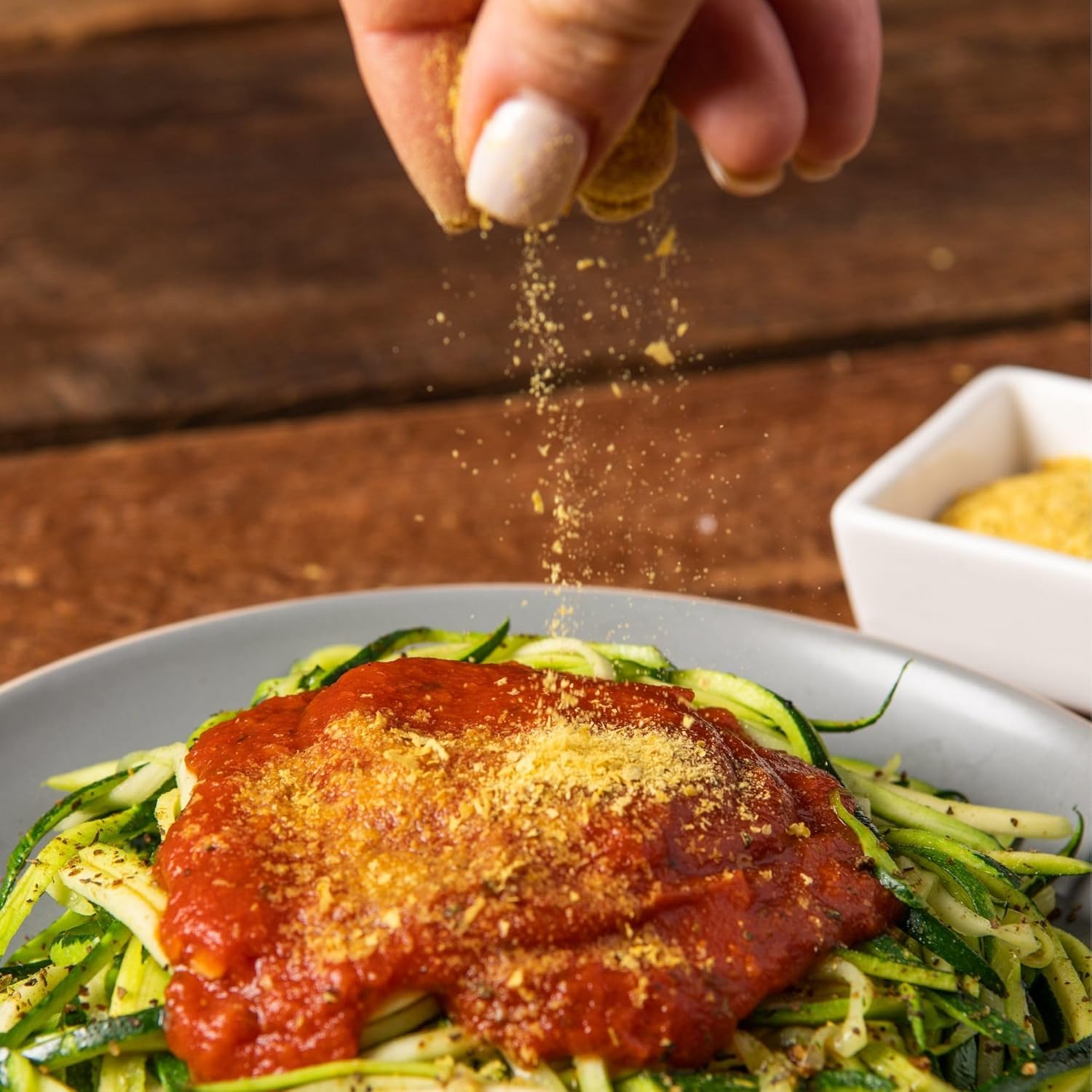 Hand sprinkling seasoning over a dish of zucchini noodles with red sauce on a wooden table.