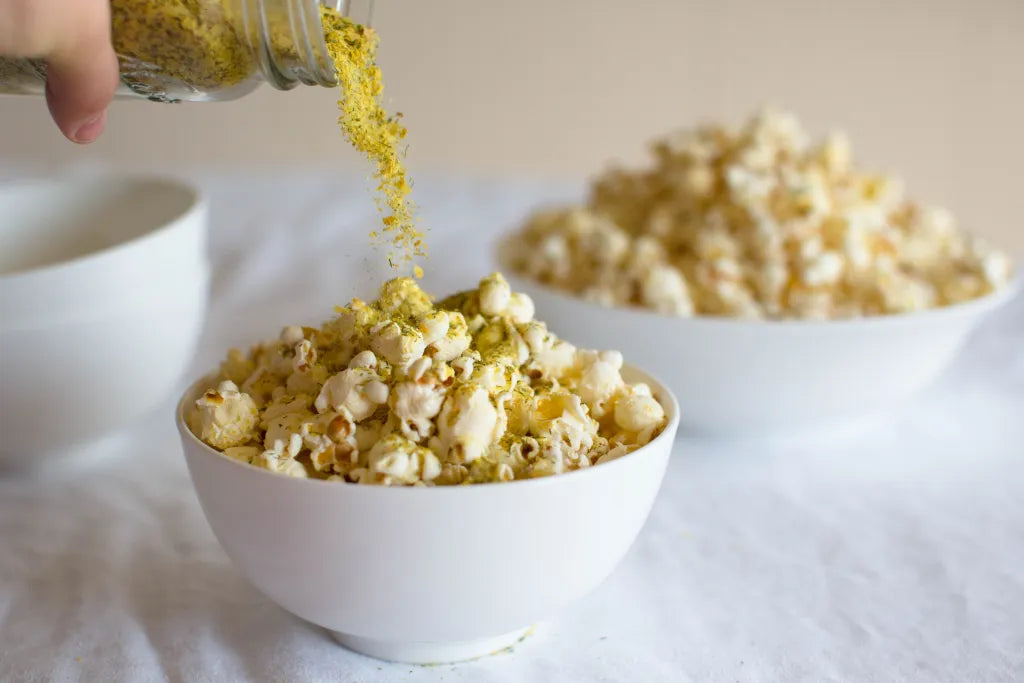 Person adding seasoning to popcorn in a bowl on a white surface